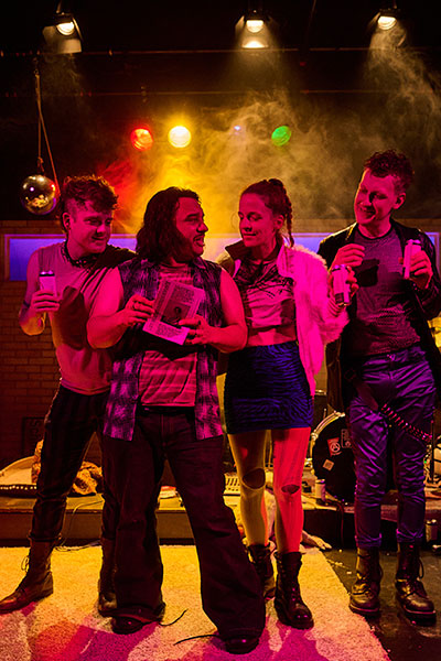 Vier junge Personen in Punk-Kleidung stehen nebeneinander in einem vernebelten Raum, der von buntem Licht erfüllt wird.