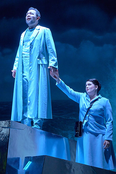 Ein Mann in hellen Farben gekleidet steht auf einer Erhöhung und singt mit Blick nach vorne gerichtet. Eine Frau, ebenfalls in helle Farben gekleidet, steht hinter und unter ihm in nimmt seine Hand.