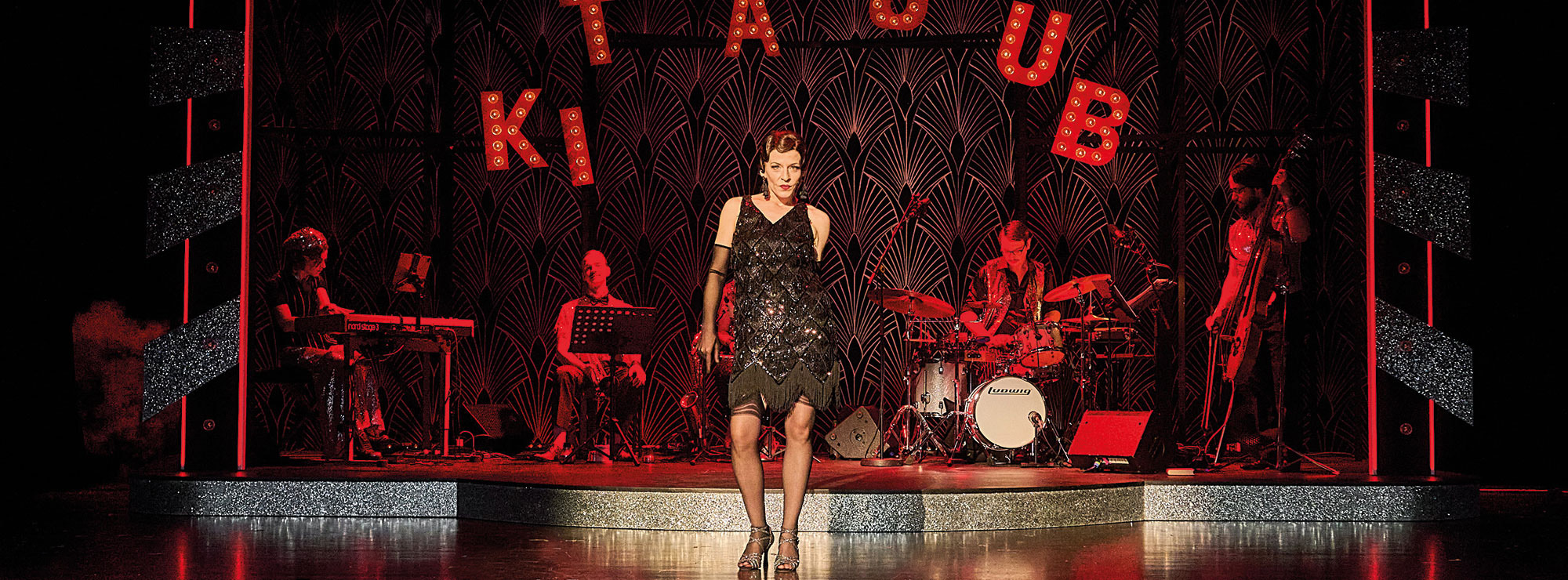 Im Hintergrund steht in großen leuchtenden Buchstaben KIT KAT CLUB, davor spielt eine Band und es tanzt eine Frau in einem glitzernden schwarzen Kleid im Stil der 1920er-Jahre.