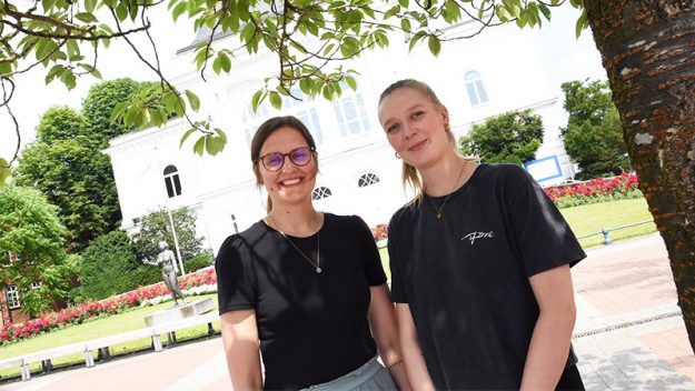 Zwei junge Frauen in schwarzen T-Shirts stehen nebeneinander vor dem Rendsburger Stadttheater und lächeln in die Kamera.